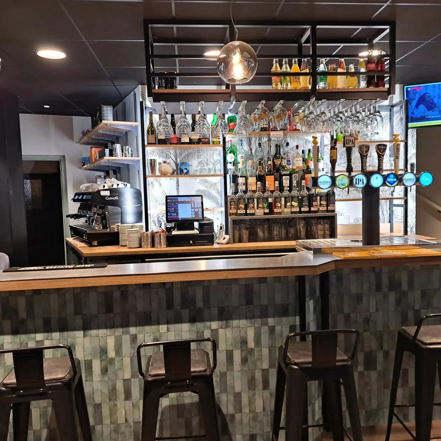L'arrière du bar : machine à café, étagères de bouteilles et de sirops, verres suspendus au-dessus du comptoir.